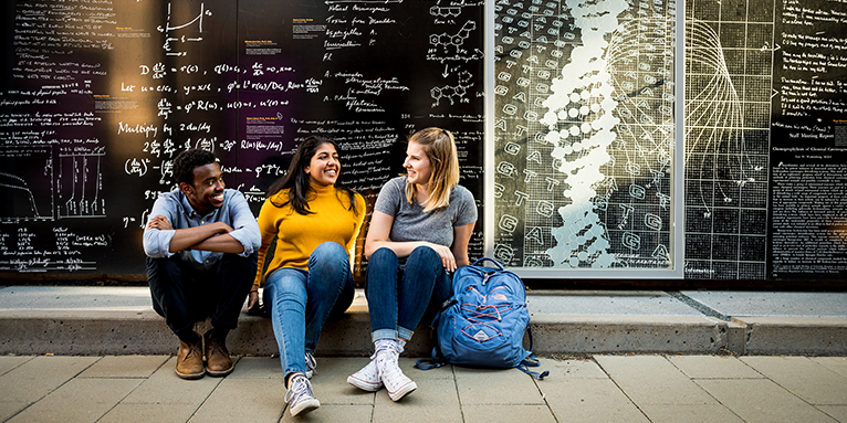 Three students sit in front of a black wall with science calculations.