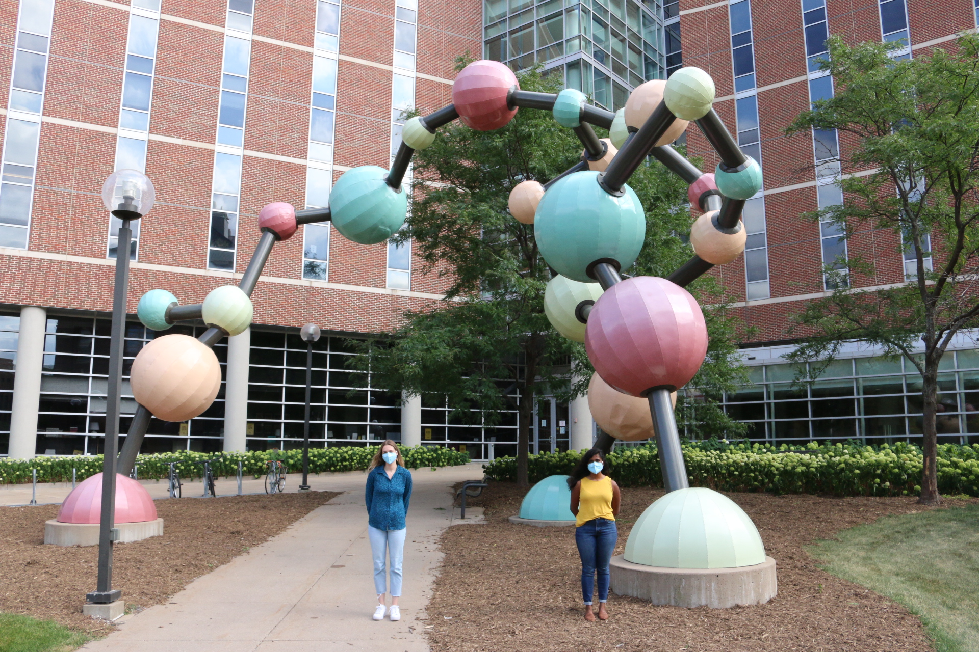 Abbey Robinson and Judee Sharon stand outside MCB wearing masks with the molecule behind them.