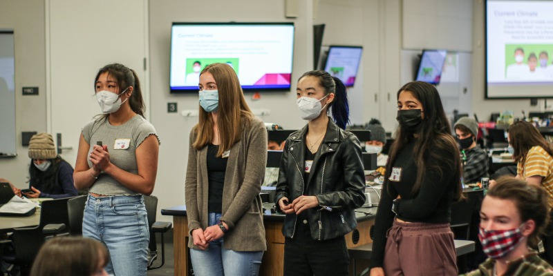 Four students present their case study solution to other students at the event. The four students stand in front of other students and presentation screens in a large classroom. 