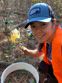 Sara sits in the woods holding a tweezers. A gallon jug of mustard solution is in the background. 