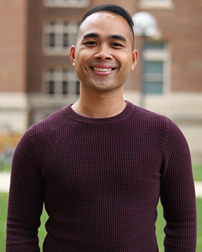 Bory poses with a smile and is wearing a maroon sweater in front of a brick building