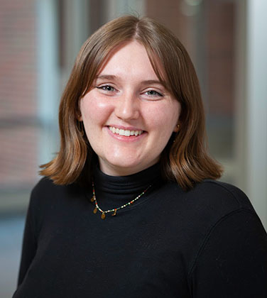 Rachel has light brown shoulder-length hair and wears a black turtleneck and necklace.