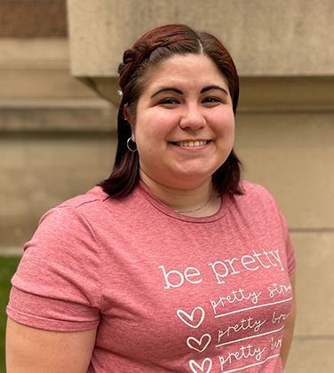 Kay has dark brown and red hair with a french braid on left side. She is wearing a pink shirt that says "be pretty"